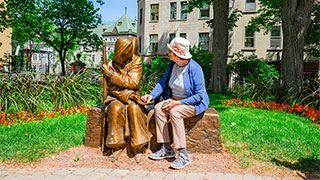 A woman in a blue cardigan and sun hat sits next to a bronze statue during an Old City Quebec walking tour.