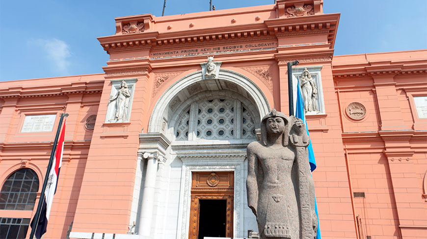 The salmon-colored exterior of the Egyptian Museum features an arched entrance and a large ancient stone statue of a pharaoh in the foreground.