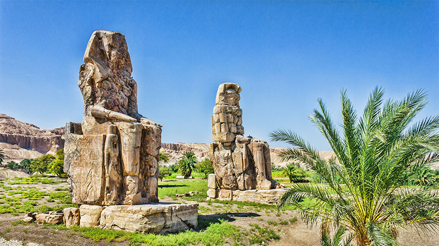 Two giant ancient stone statues sit in a grassy field near Luxor's Valley of the Kings with mountains in the background.