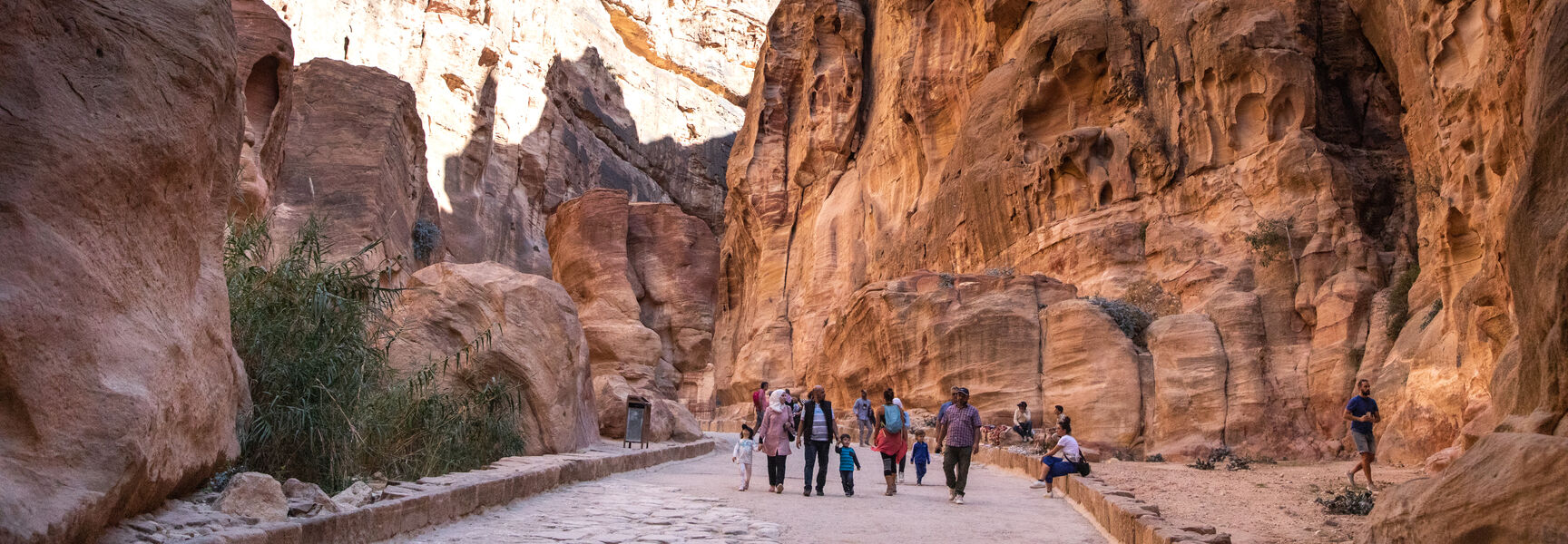 Tourists walking along a narrow stone path between towering red sandstone cliffs during a walking tour in Petra.
