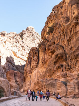 Tourists walking along a narrow stone path between towering red sandstone cliffs during a walking tour in Petra.