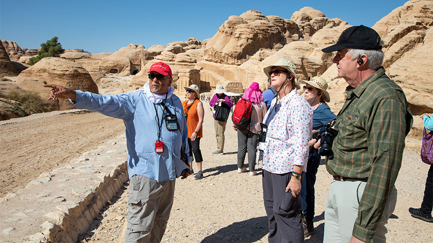 A tour guide in a red hat points toward sandstone rock formations for a group of tourists on a Petra walking tour.