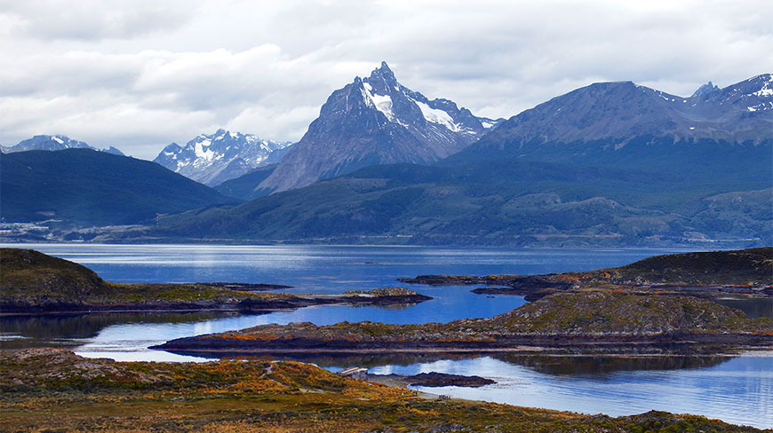 Snow-capped mountains tower over the blue waters and rocky shorelines of Tierra del Fuego National Park under a cloudy sky.