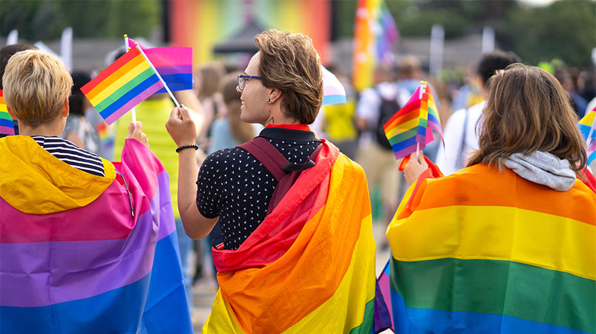 People celebrate at a pride parade, holding rainbow and bisexual flags while draped in colorful pride flag capes.