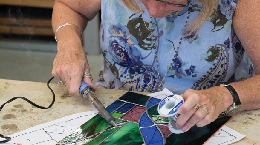 A person at Snow Farm uses a soldering iron to carefully join colorful pieces of stained glass together on a workshop table.