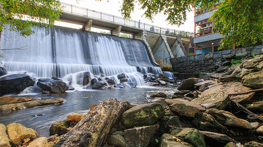 Water cascades over a concrete dam at the Montreat Conference Center in North Carolina, with a bridge above and a rocky stream below.