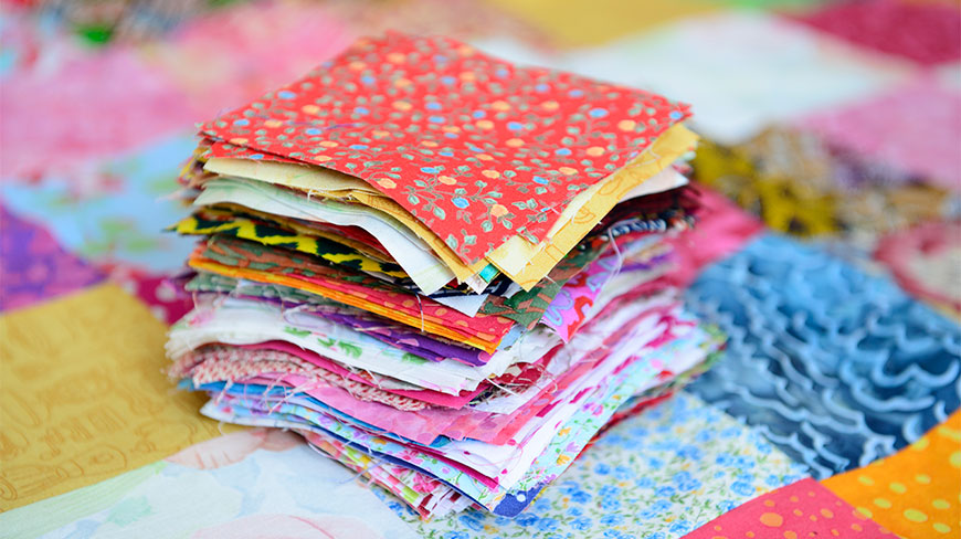A tall stack of colorful patterned quilt fabric squares sits atop a finished patchwork quilt.