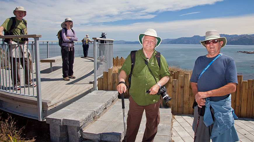 NZ participants in hiking gear and hats stand on a coastal lookout overlooking a scenic bay and mountains in New Zealand.