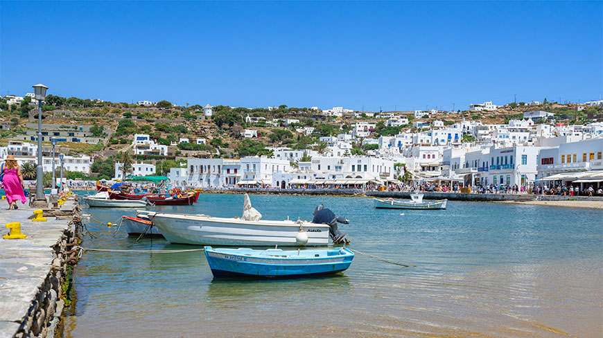 A scenic view of the Mykonos harbor with traditional white buildings on the hillside and small boats floating in the clear blue water.
