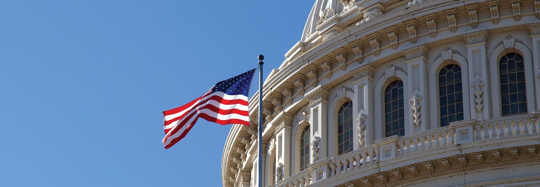 The American flag waves in front of the U.S. Capitol dome against a clear blue sky in Washington, D.C.