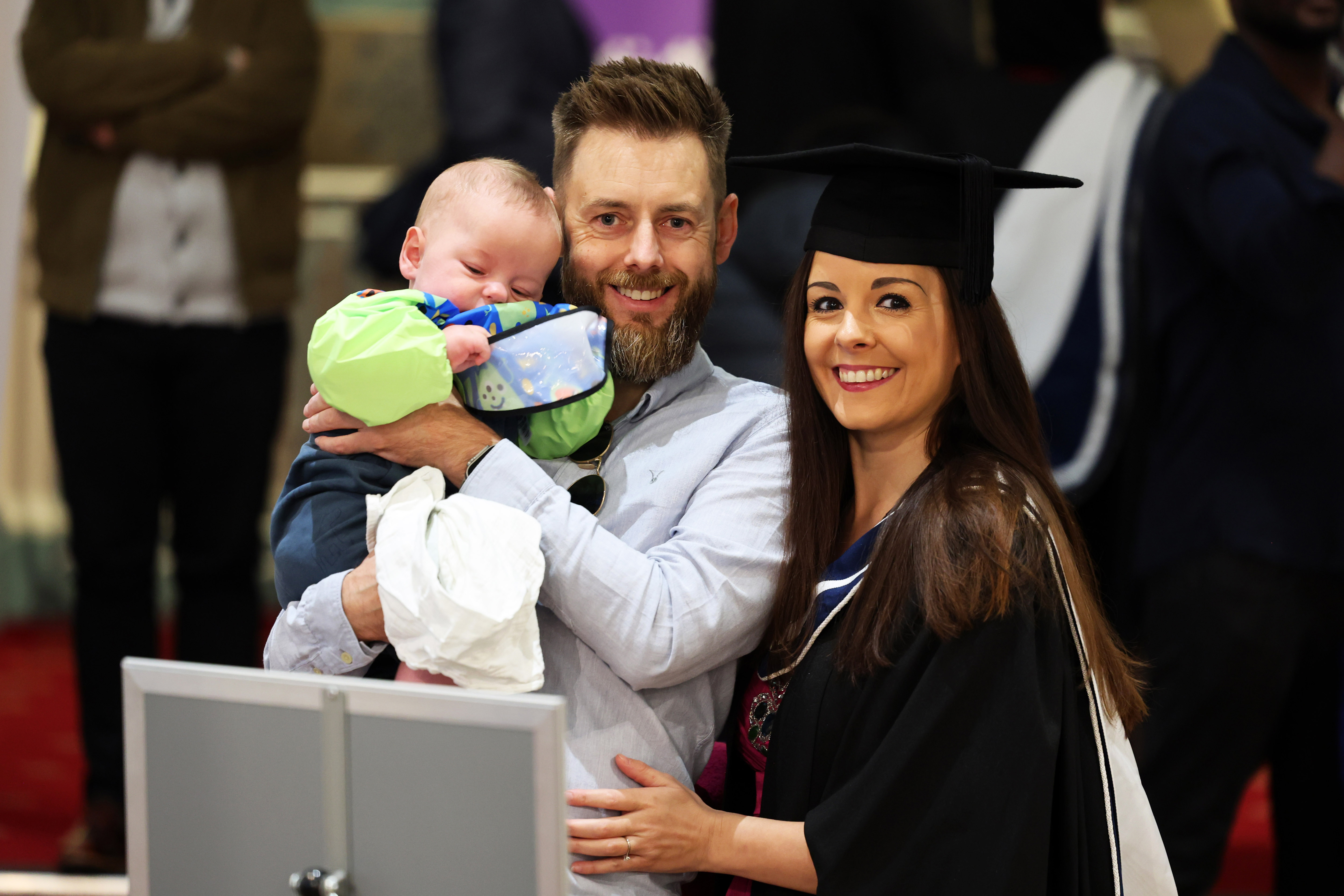 Female graduate in cap and gown posing with her partner and toddler child