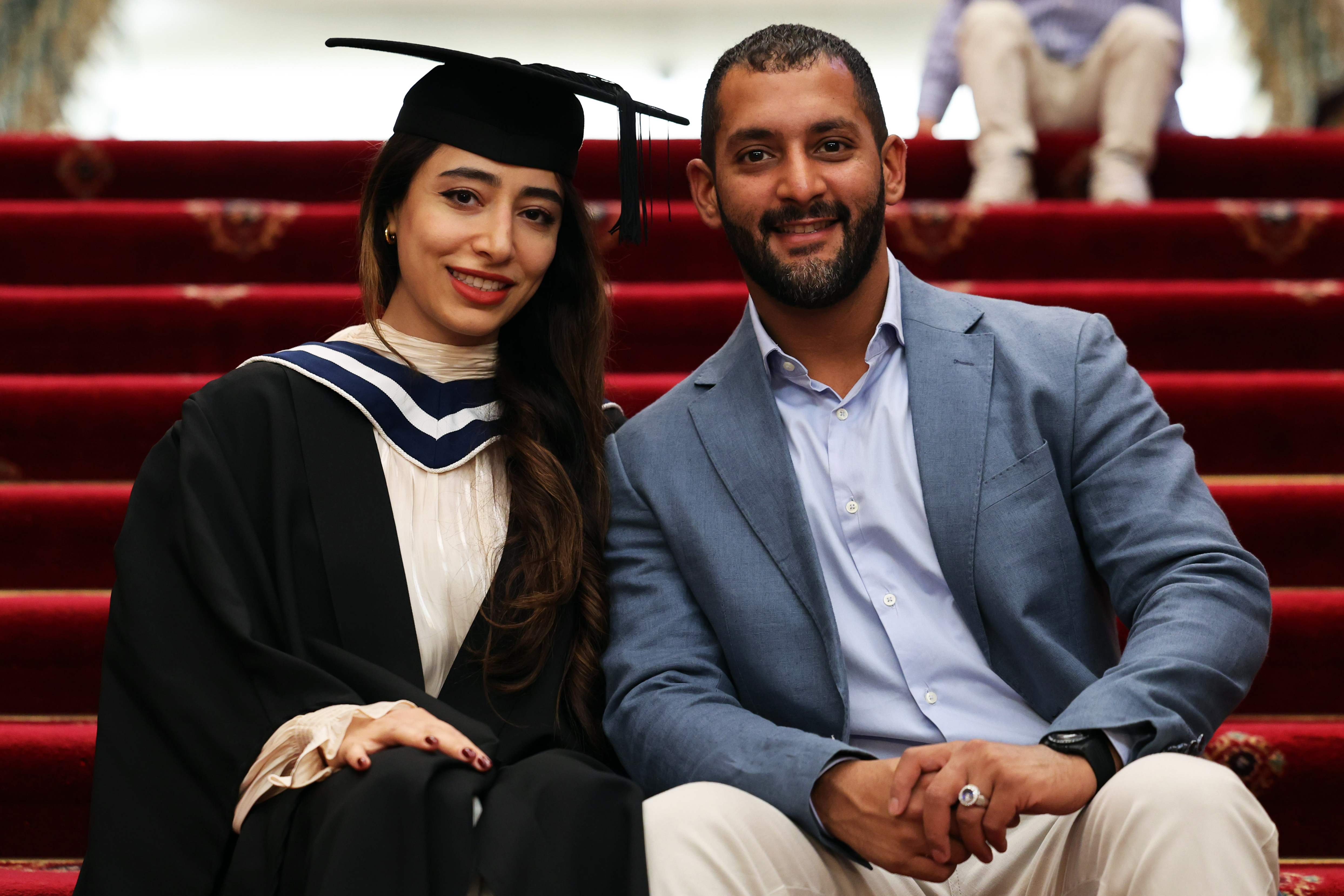 Female graduate in cap and gown posing with a man (boyfriend, husband or loved one) after ceremony, seated on stairs