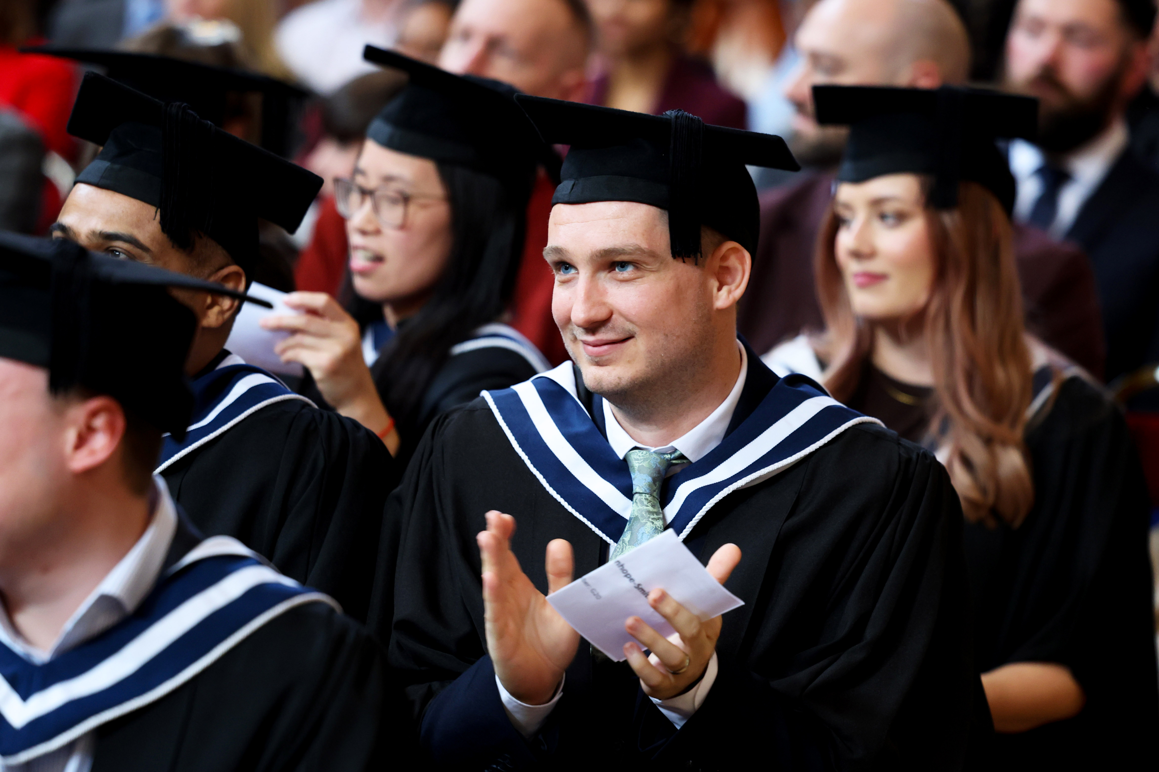 Group of graduates in cap and gown, focus is on one male graduate who is clapping his hands