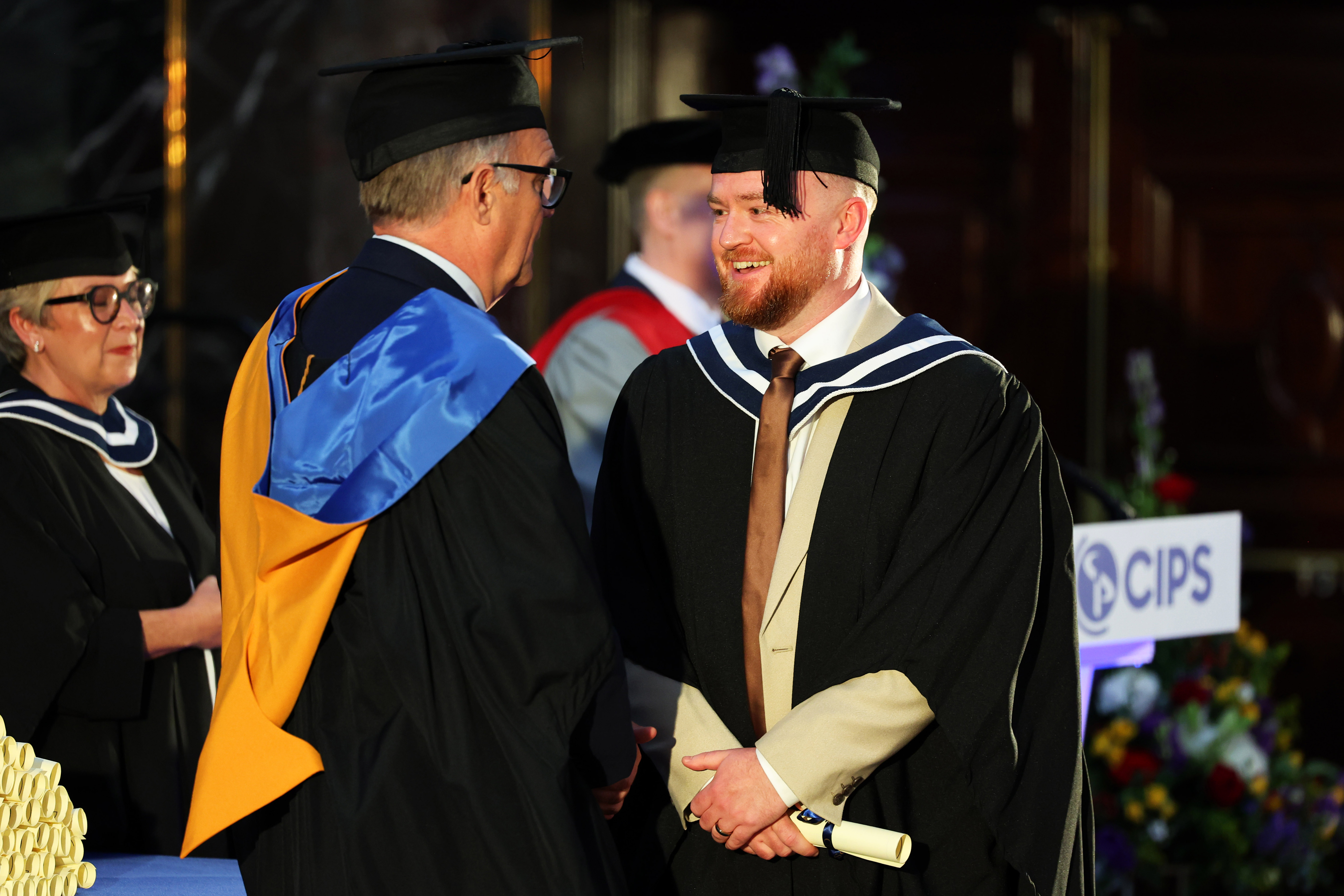 Male graduate in cap and gown on the stage, smiling as he receives his diploma from CIPS CEO