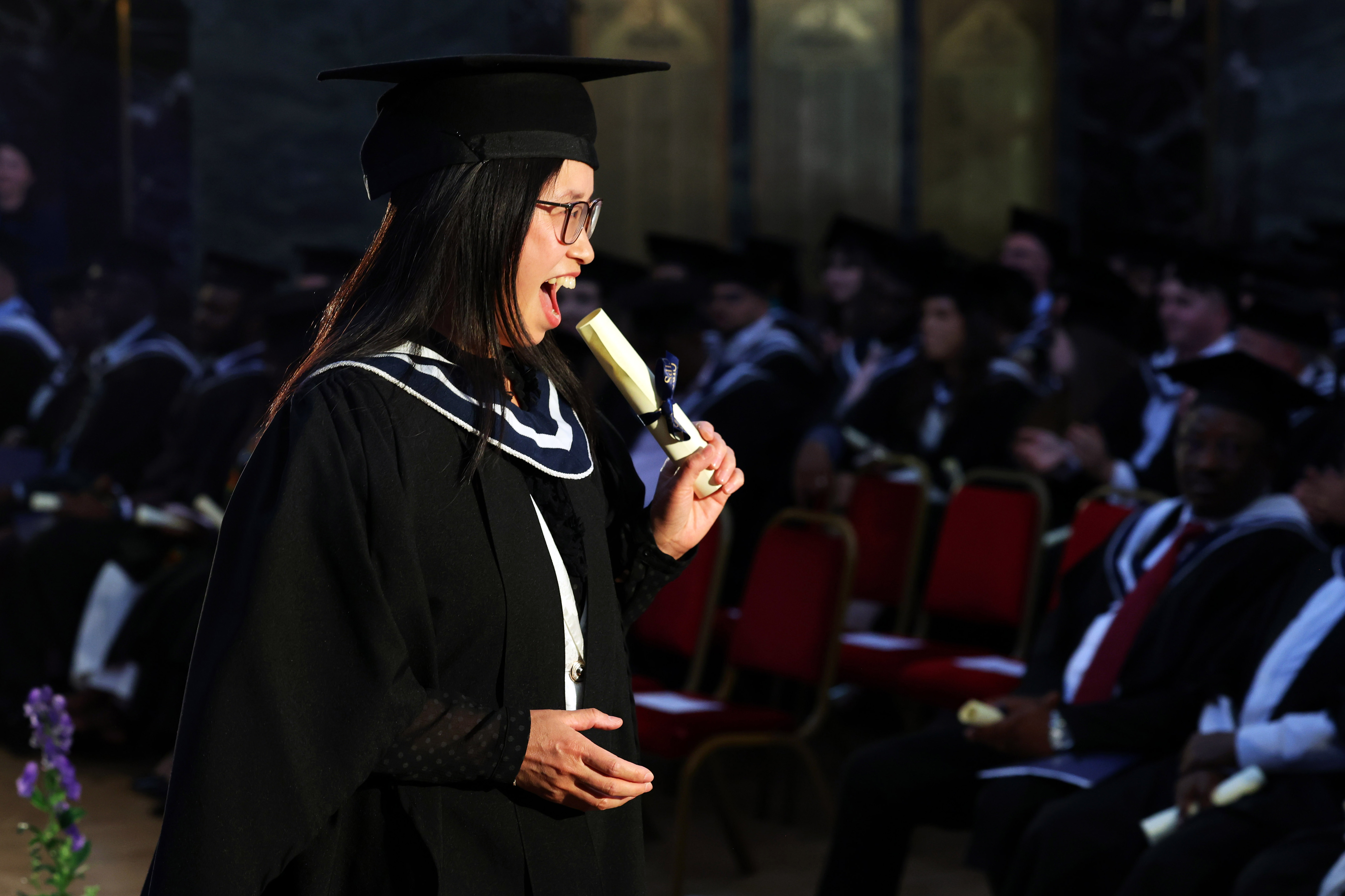 Female graduate in cap and gown on the stage holding her diploma as she walks back to audience