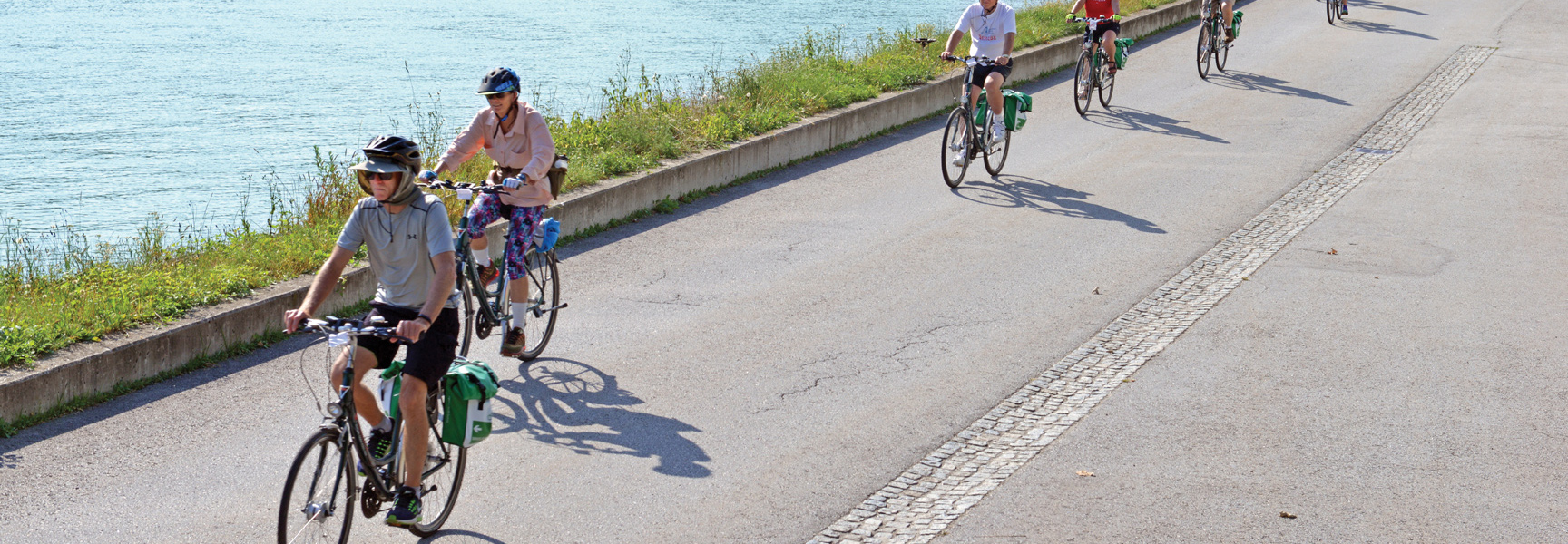 A group of cyclists ride on a paved path alongside the Danube River in Austria and Germany on a sunny day.
