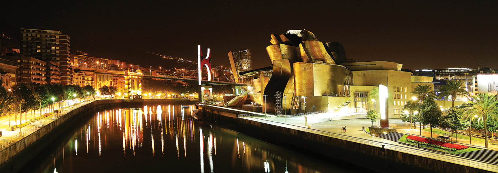 The Guggenheim Museum in Bilbao, Spain, is lit up at night, with city lights reflecting in the Nervión River.