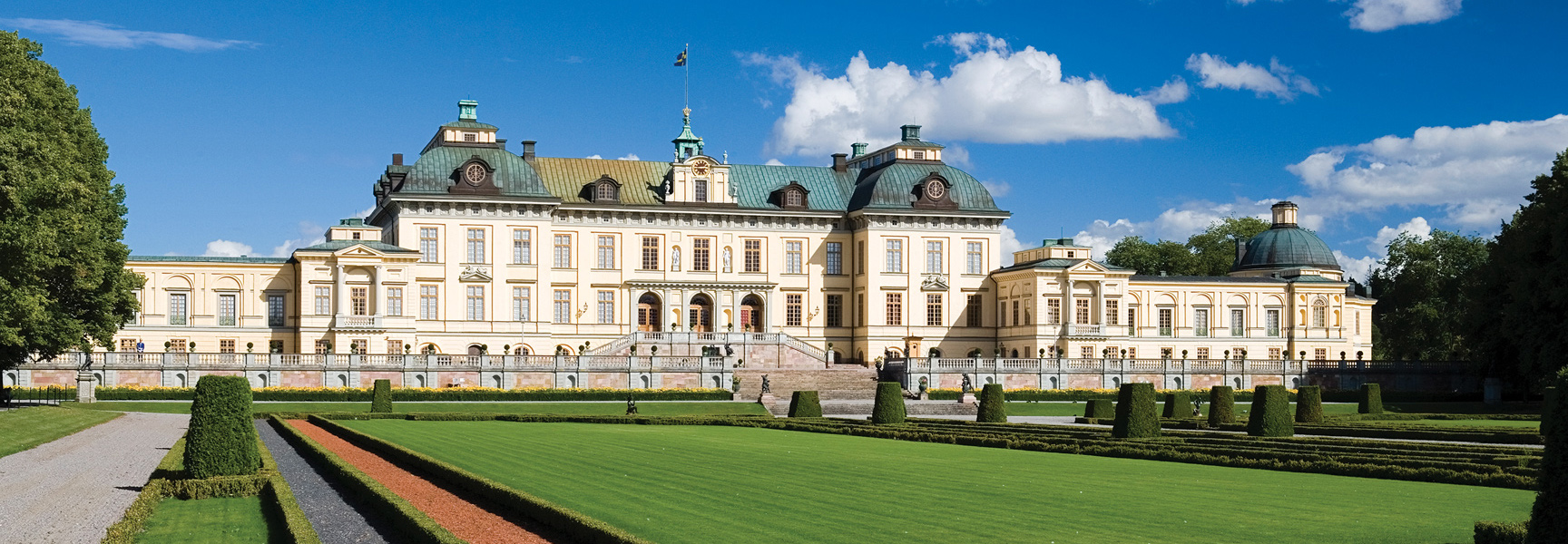 The grand Drottningholm Palace in Sweden, with its manicured gardens and sprawling lawn under a bright blue sky.