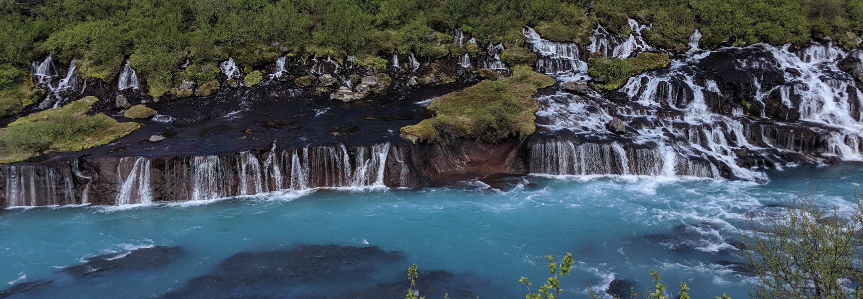A wide series of waterfalls cascade over dark lava rock into a vibrant, turquoise river in Iceland.