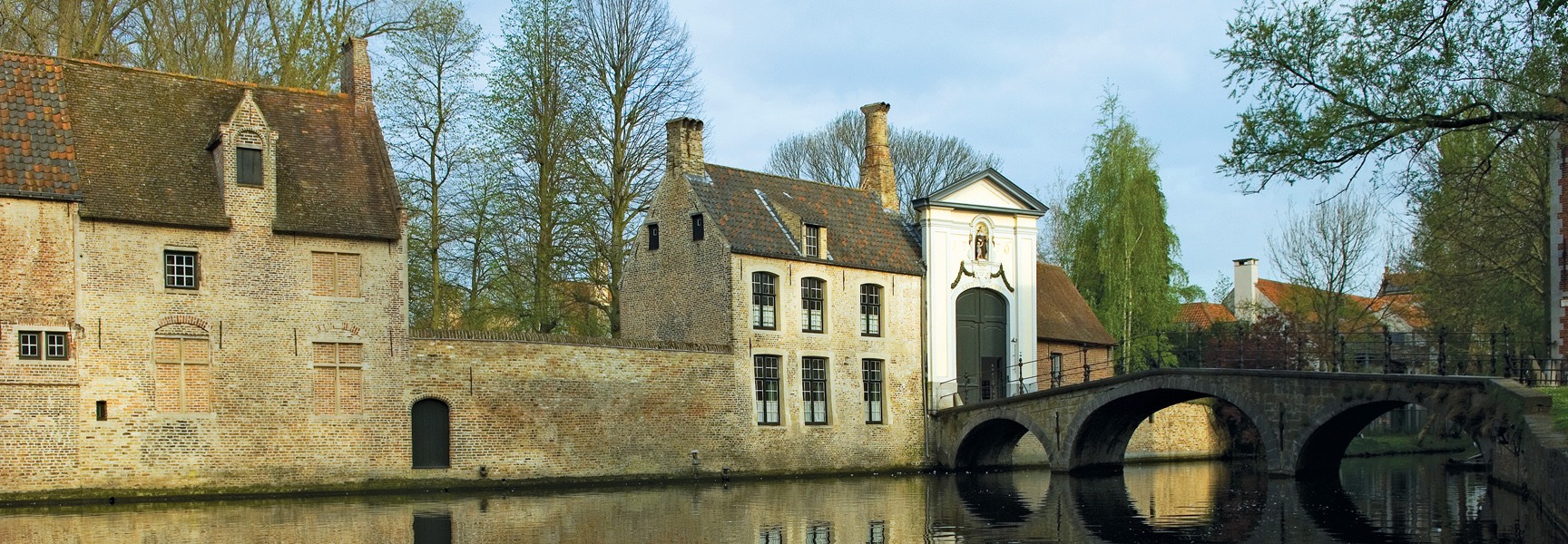 Old brick buildings and an arched stone bridge along a canal in the Netherlands or Belgium.