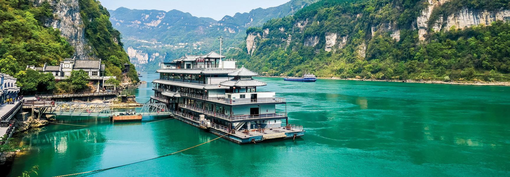 A large riverboat with traditional Chinese architecture is docked on the bright green Yangtze River in China, surrounded by steep, verdant mountains.
