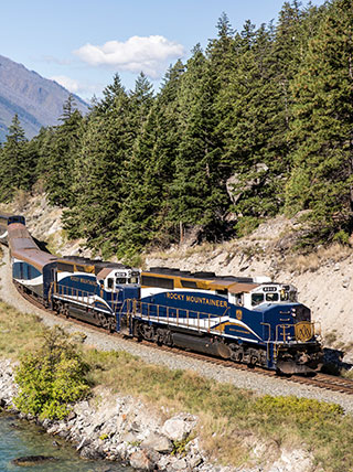 The blue and gold Rocky Mountaineer train travels on a track beside a forested mountain and waterway in Alberta/British Columbia.
