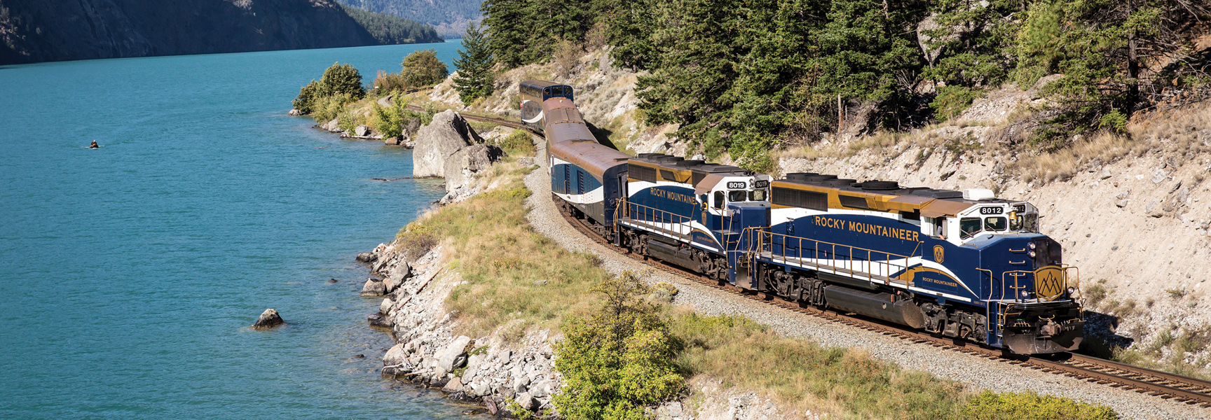 The Rocky Mountaineer train travels alongside a turquoise lake surrounded by forested mountains in Alberta/British Columbia.