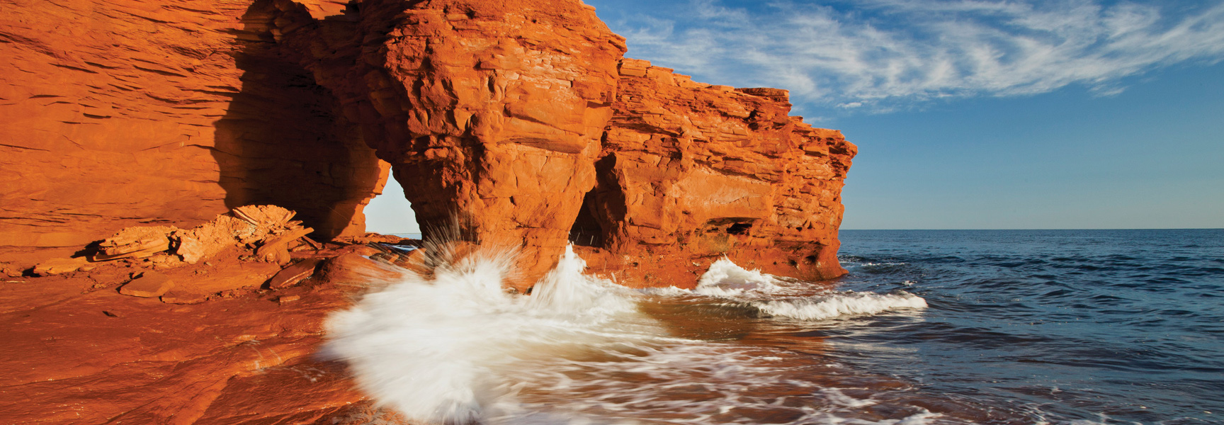 Waves crash against the iconic red sandstone cliffs of Prince Edward Island under a blue sky.