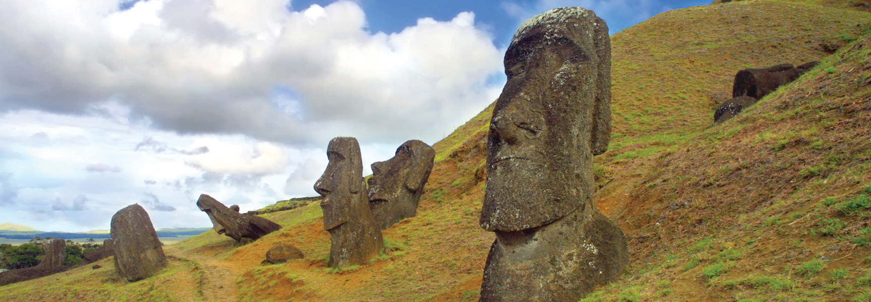Several large moai statues stand on a grassy hillside under a partly cloudy sky on Rapa Nui, Chile.