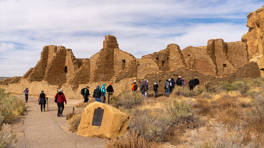 A group of people on a guided tour walk past the large, ancient stone ruins of Chaco Canyon in New Mexico.