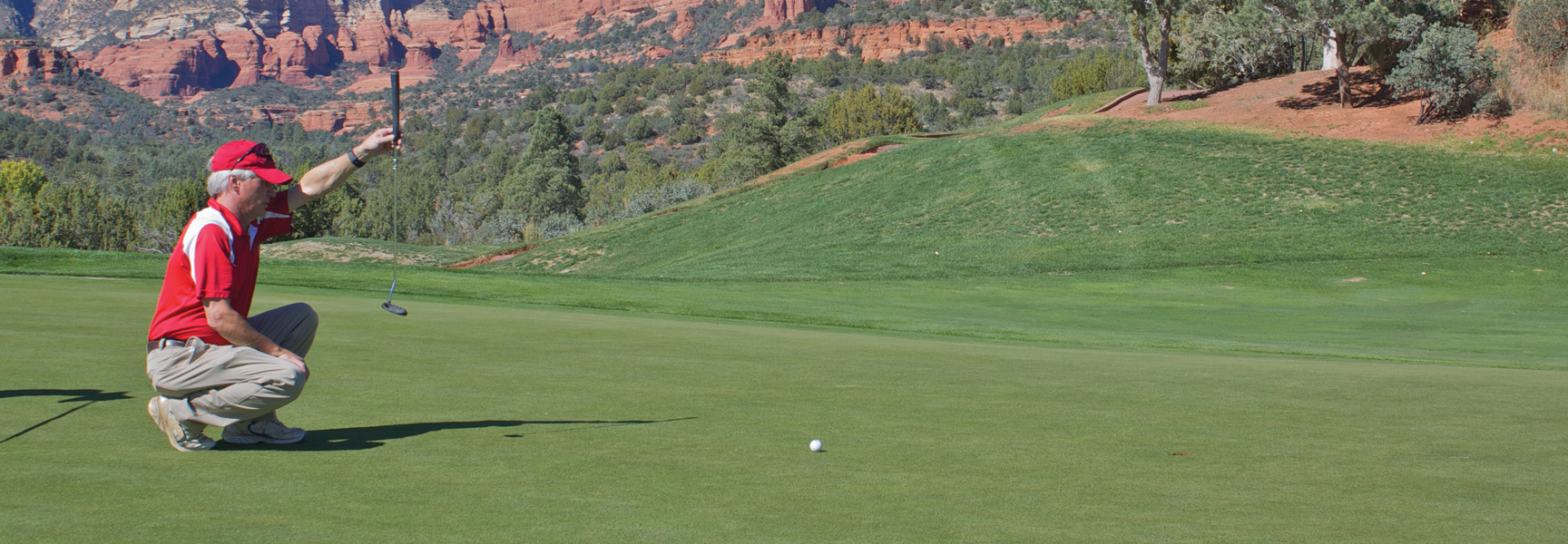 A man lines up a golf putt on a lush green with the red rocks of Sedona, Arizona, visible in the distance.
