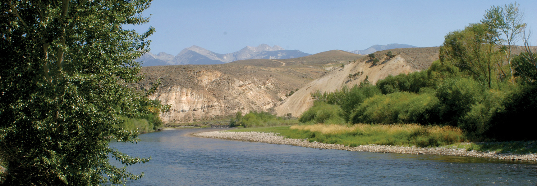 A scenic view of the Lower Salmon River in Idaho shows it winding past tree-lined banks with arid hills and mountains behind.