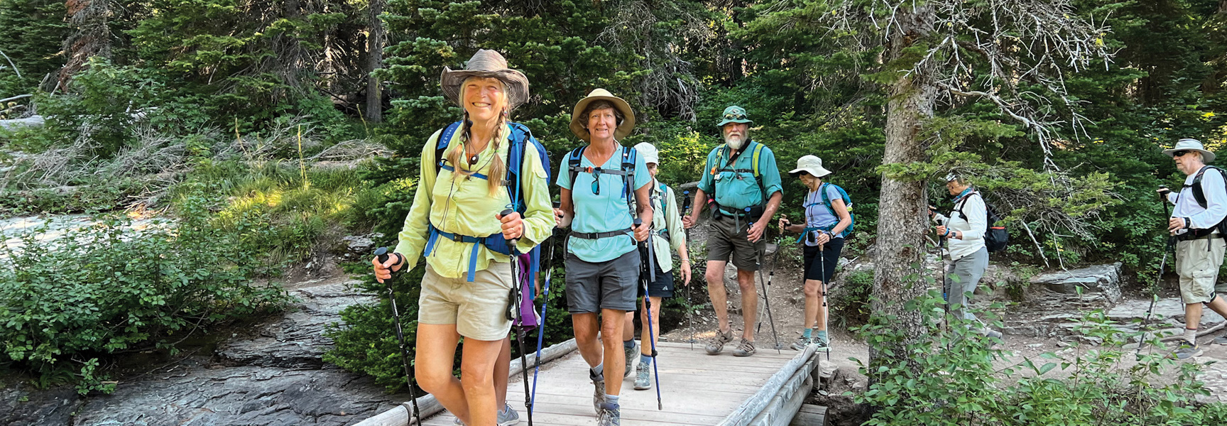 A group of hikers cross a wooden bridge on a forested trail in Glacier National Park, Montana.