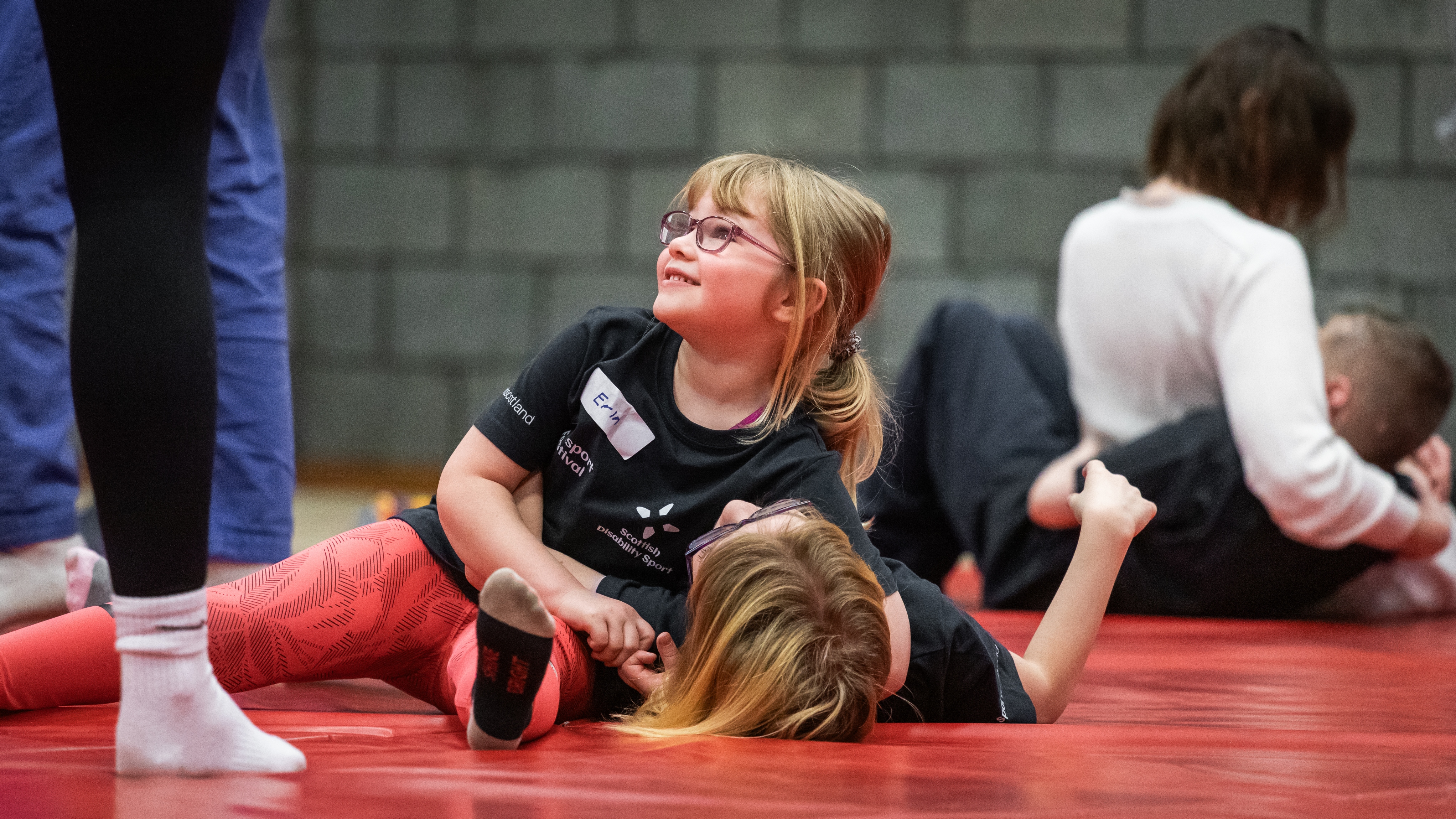 A young girl wearing glasses smiles while playfully participating in a sports activity on a red mat, surrounded by other children and adults in a gym setting.