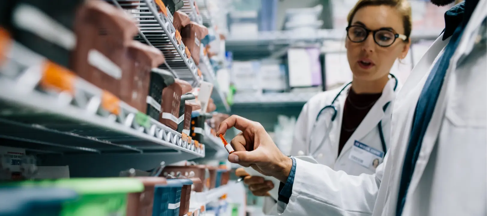 
A low-angle close-up shot of two people in a pharmacy or medical storage area.

The person in the foreground, wearing a white lab coat, is holding up a small orange and white capsule or pill to examine it. The second person, also in a white lab coat and wearing glasses and a stethoscope, is looking at the pill and appears to be speaking or discussing it. They are surrounded by metal shelving racks filled with various labeled drawers and bins.
