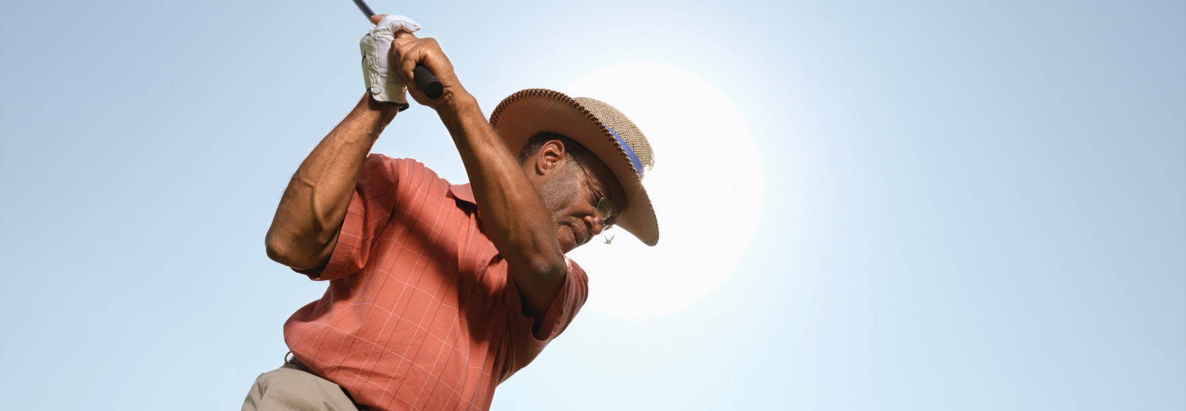 An older man wearing a sun hat and glasses follows through on his golf swing under the bright Utah sun.