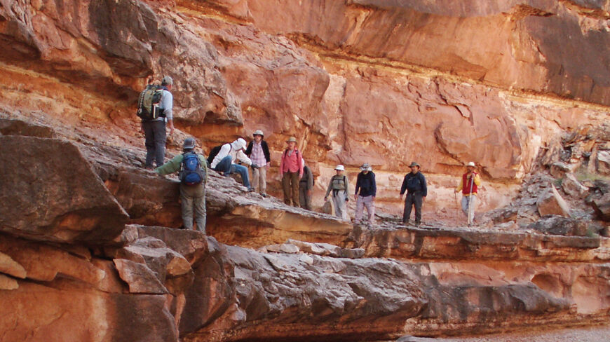 A group of hikers with backpacks navigate a steep rocky ledge within the red rock vistas of Marble Canyon, Arizona.