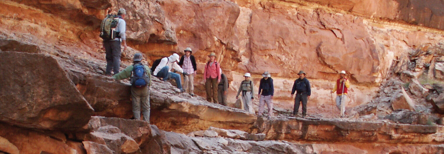 A group of hikers with backpacks navigate a steep rocky ledge within the red rock vistas of Marble Canyon, Arizona.