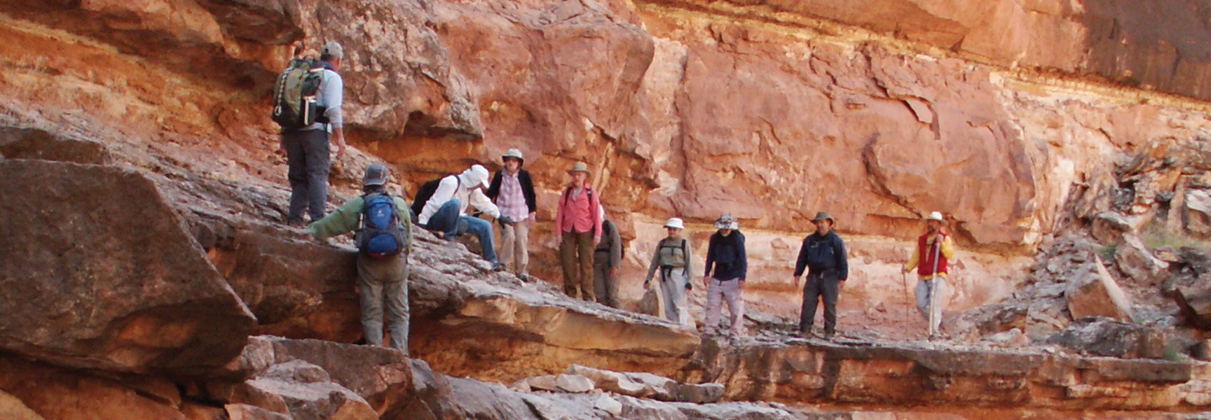 A group of hikers traverse a narrow, rocky ledge alongside a sheer red rock canyon wall in Marble Canyon, Arizona.