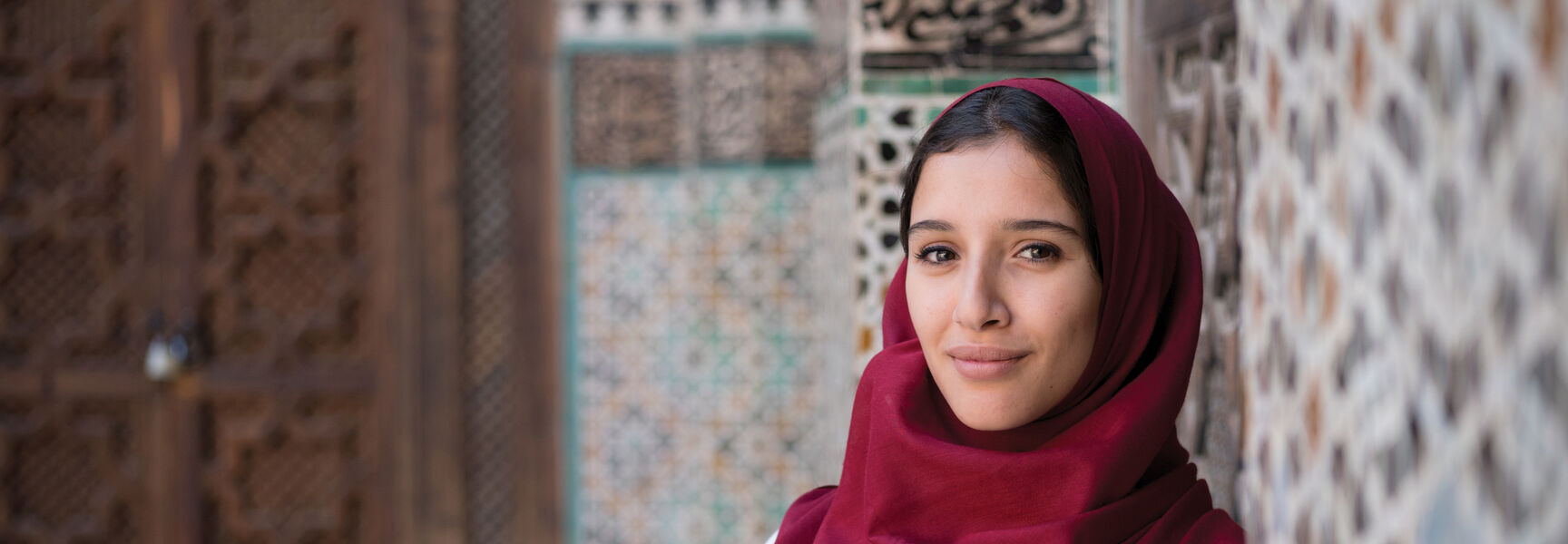 A woman in a red headscarf smiles in front of intricate mosaic tiles and carved wooden doors in Morocco.