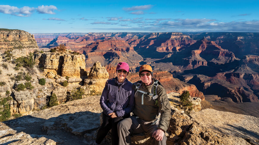 A couple in hiking gear sits on a rim overlooking the ancient landscapes of the Grand Canyon in Arizona.