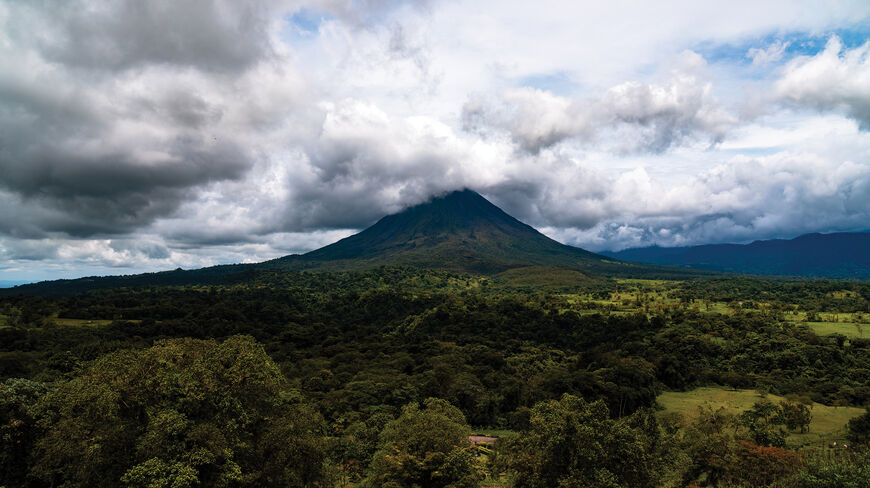 A large volcano peak partially shrouded in clouds rises above the lush rainforest and green fields of Costa Rica.