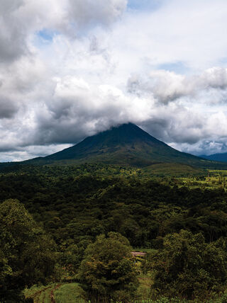 A large volcano peak partially shrouded in clouds rises above the lush rainforest and green fields of Costa Rica.