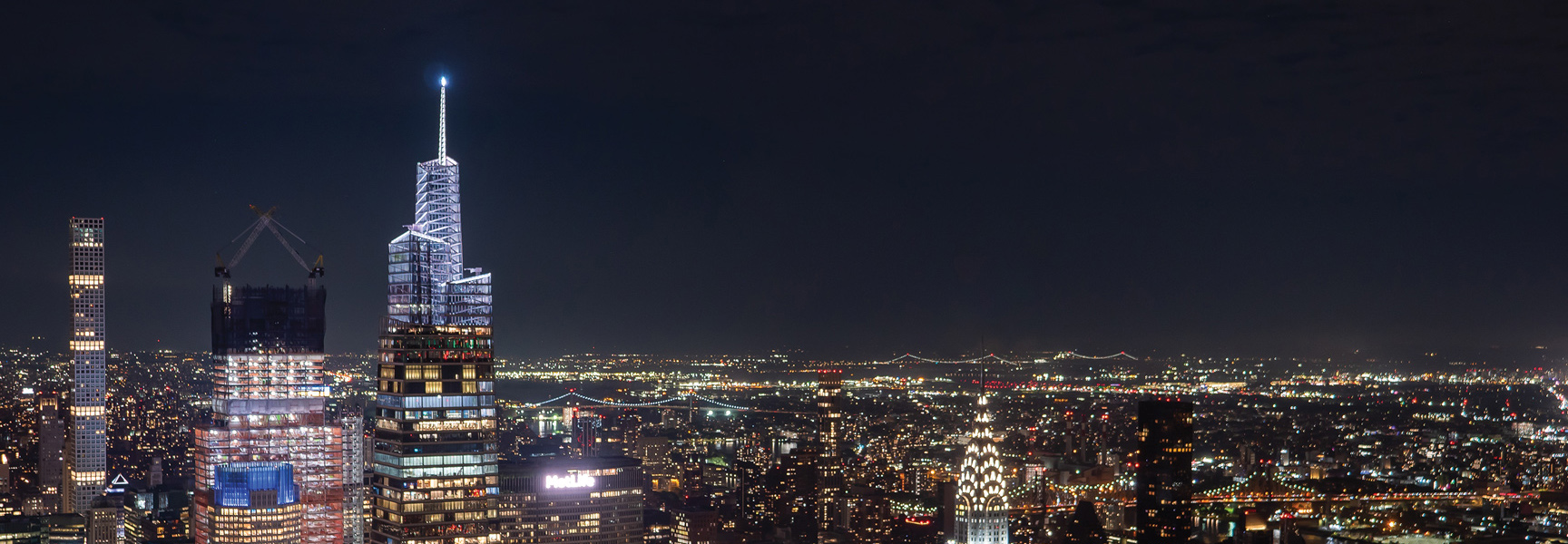 A panoramic nighttime view of the New York City skyline with illuminated skyscrapers, including one under construction, and bridges in the distance.