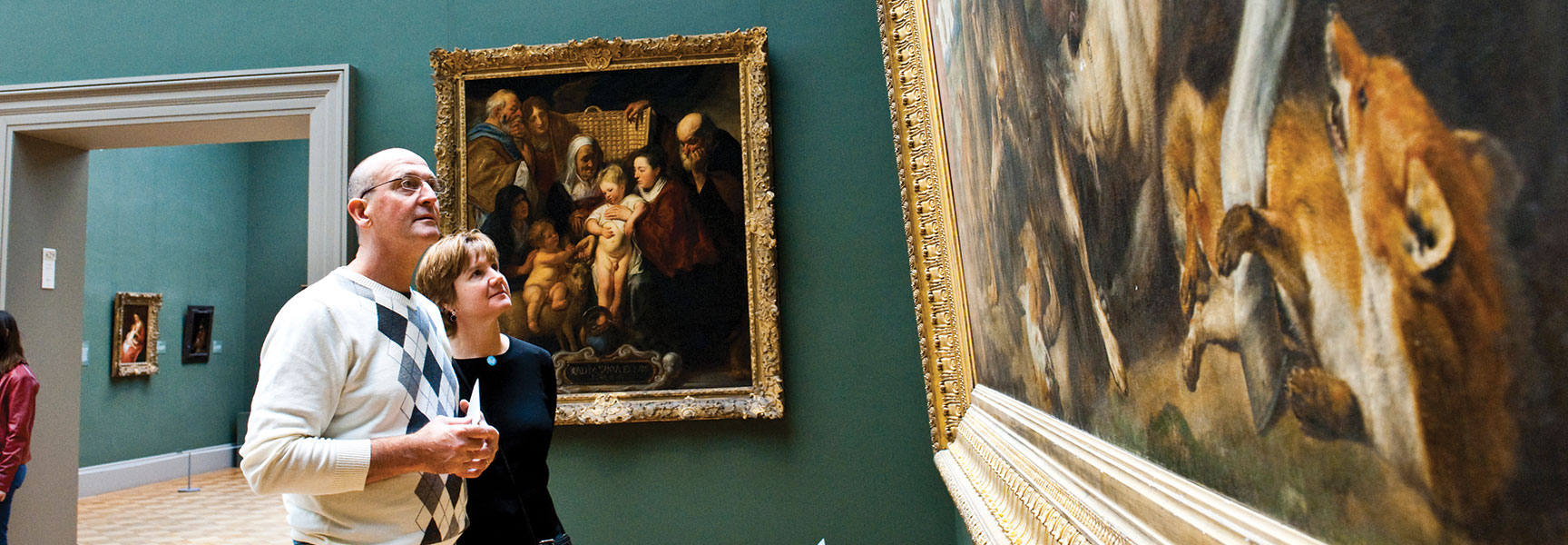 A man and a woman look up at large, ornate framed paintings on a dark green wall in a New York art gallery.