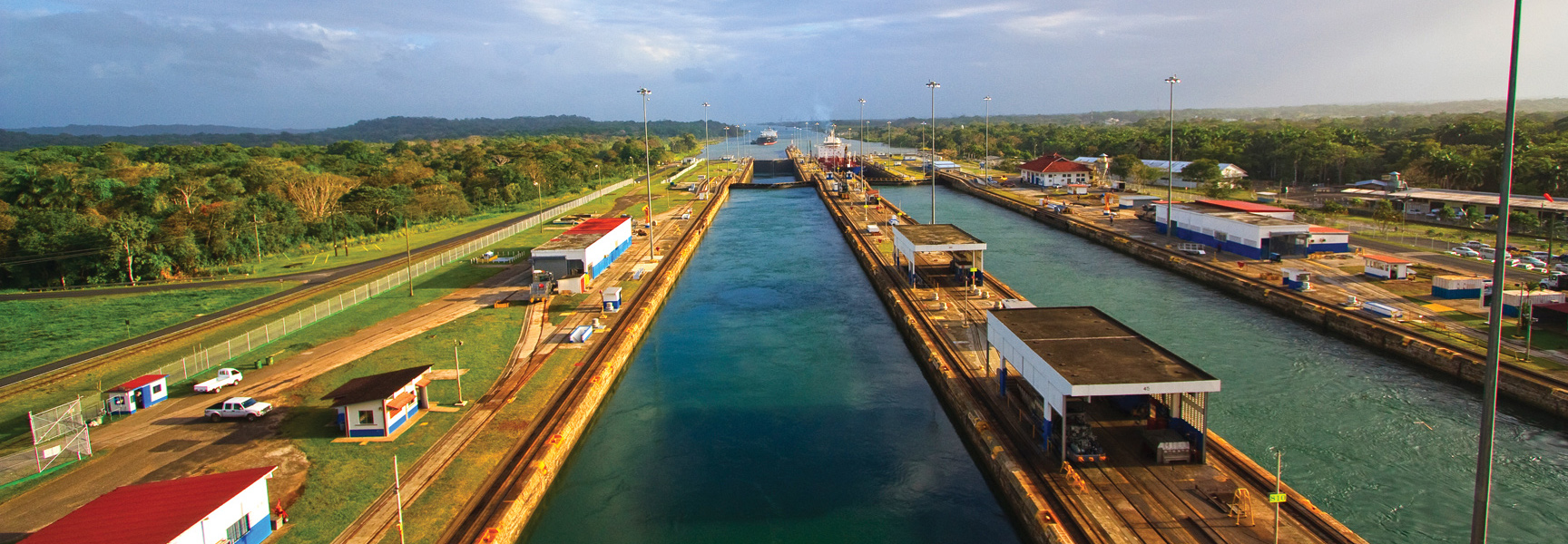 A wide, aerial view of cargo ships passing through the Panama Canal locks, surrounded by lush green jungle and industrial buildings.