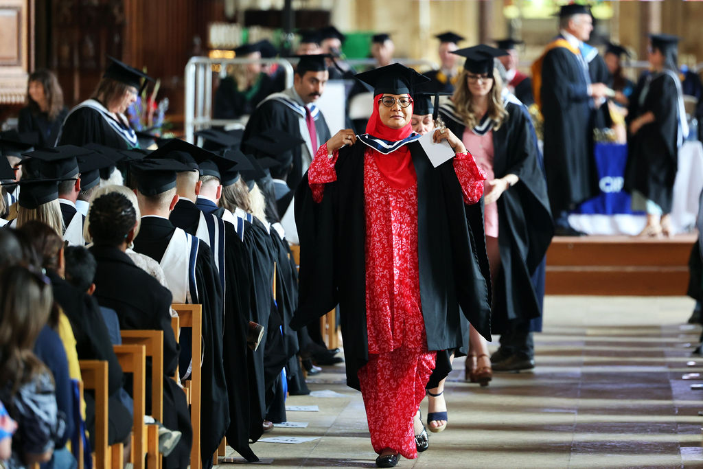 A woman in a red outfit, headscarf, and graduation gown walking down the aisle