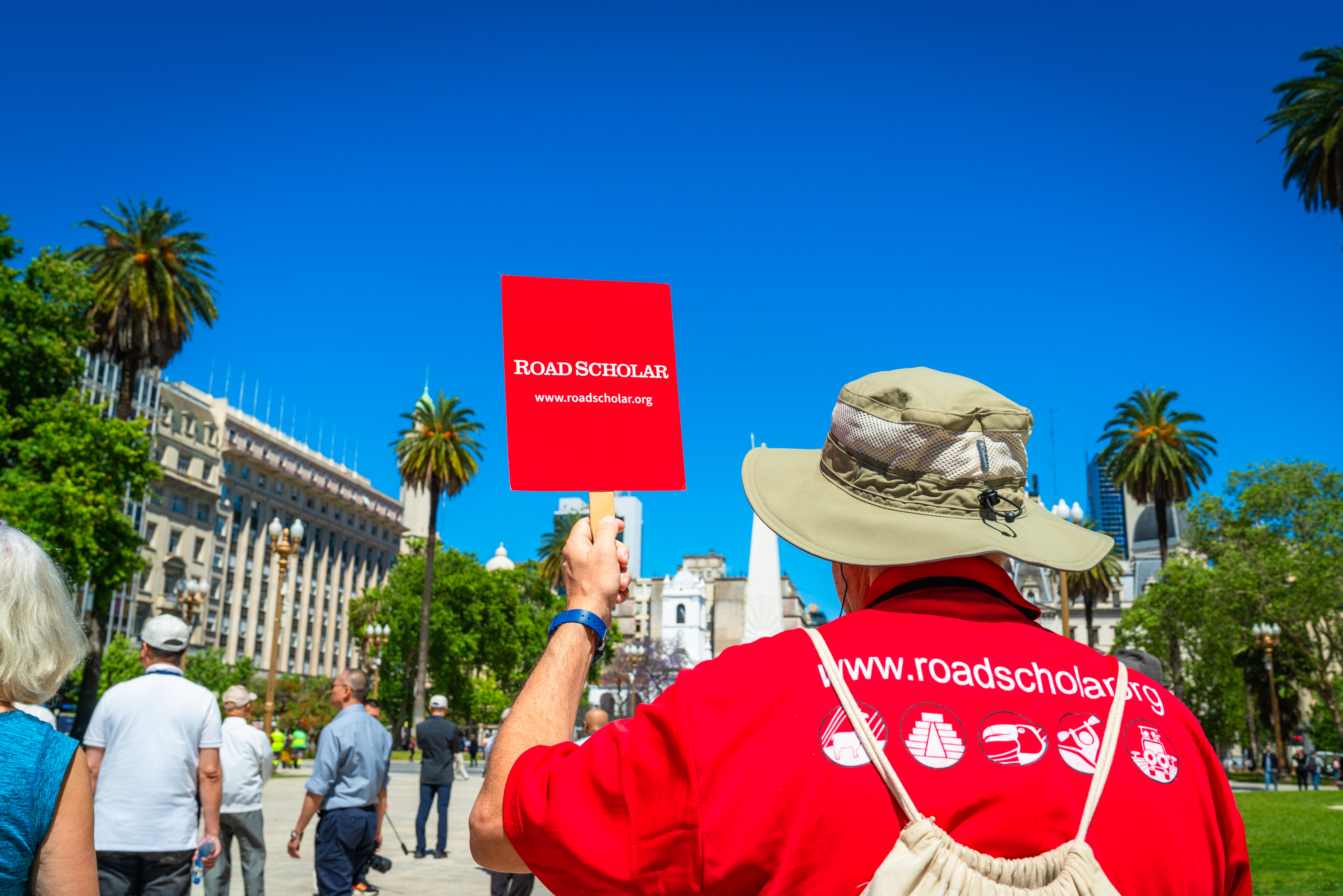 A leader in a red Road Scholar t-shirt holding a red Road Scholar sign while walking through a city