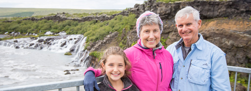 A happy young girl and her grandparents smile together for a photo in front of a scenic river and rocky landscape.