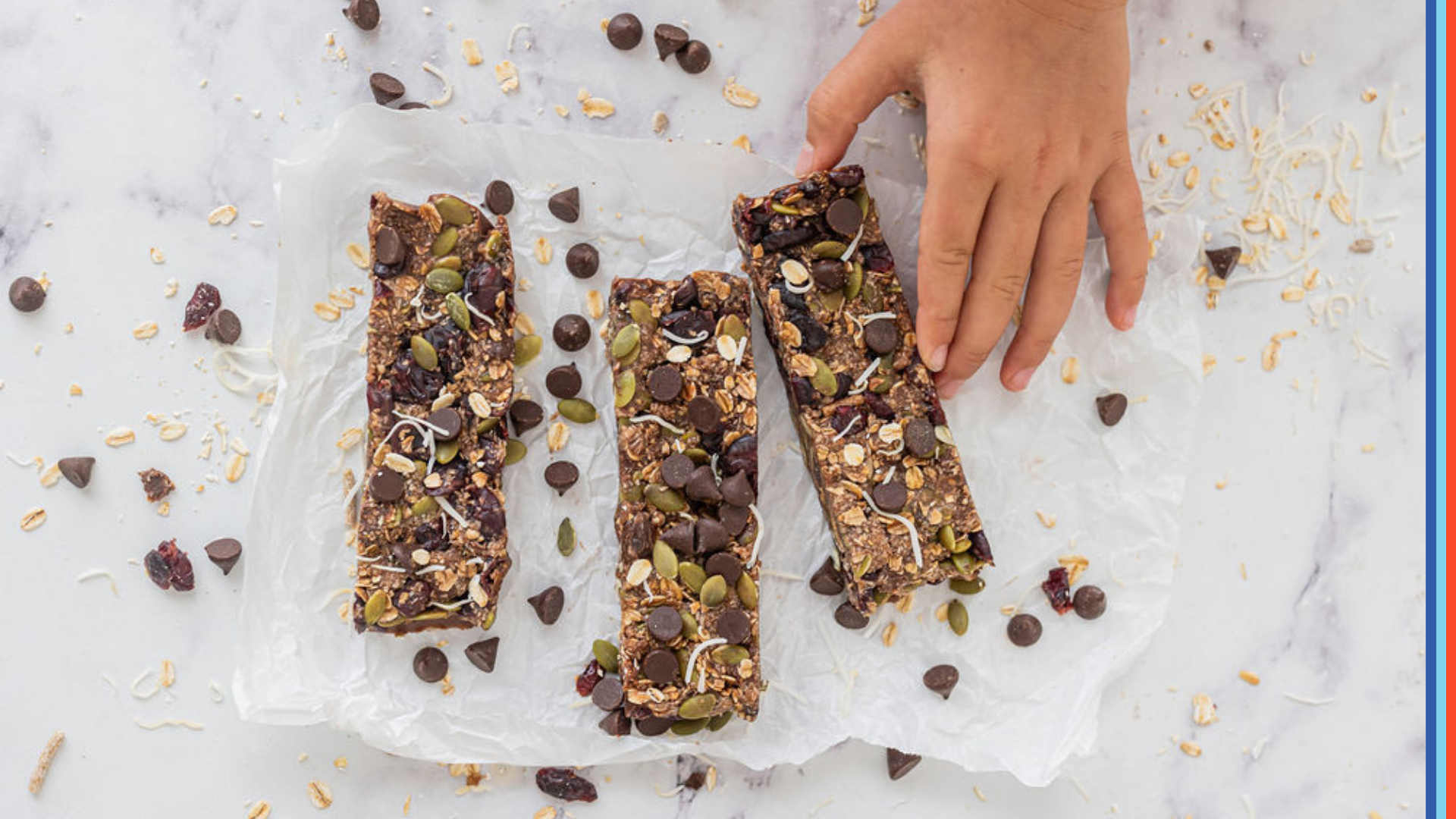 Three homemade granola bars are placed upon baking paper. The bars are sprinkled with pepitas, dark chocolate drops and shredded coconut.
